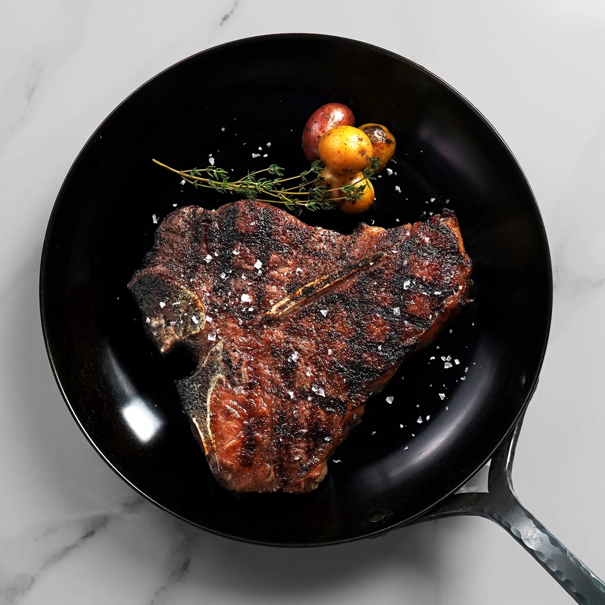 A seared T-bone steak with roasted baby potatoes and thyme in a black skillet on a marble countertop.
