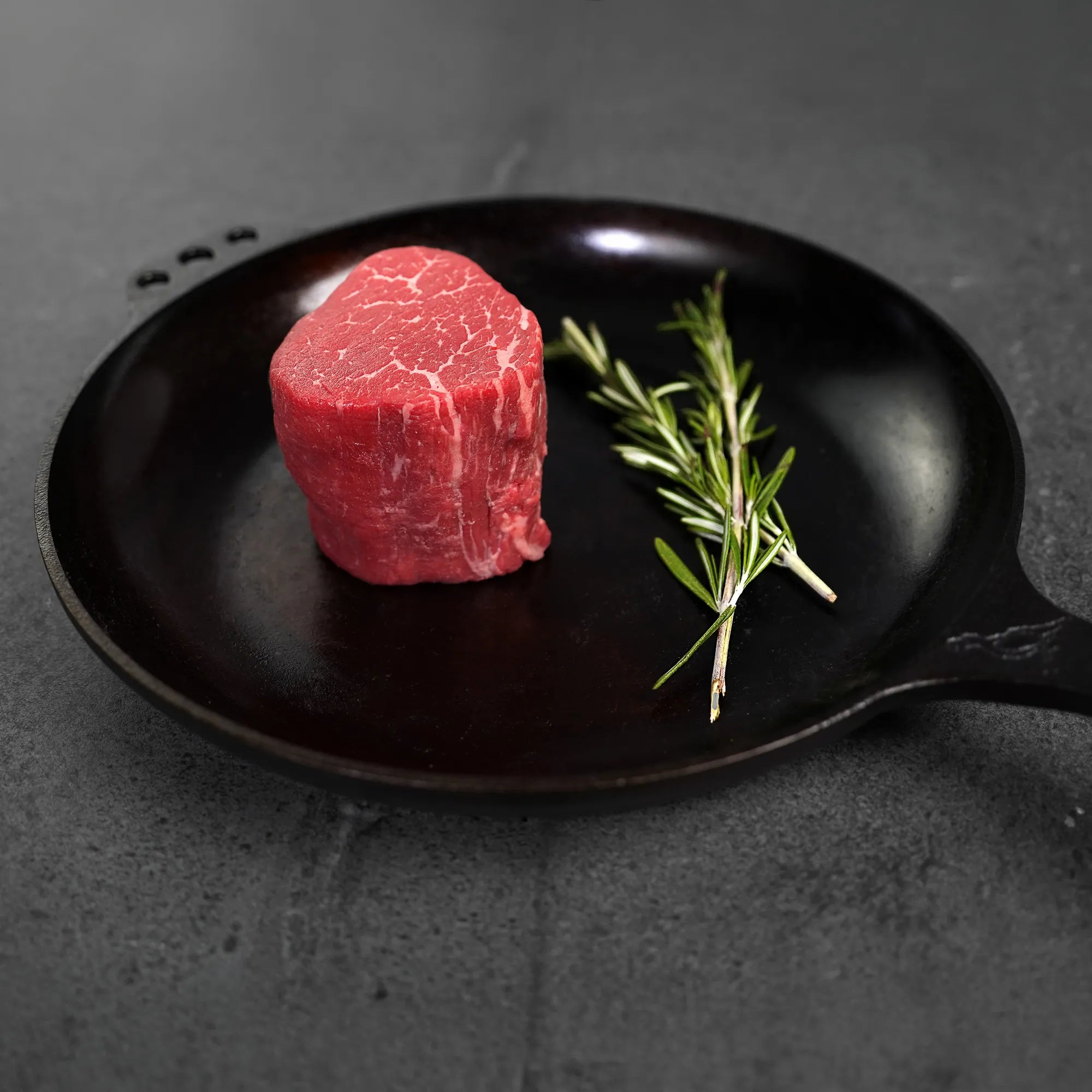 A thick-cut filet steak in a cast-iron skillet with fresh rosemary sprigs, resting on a dark stone surface.
