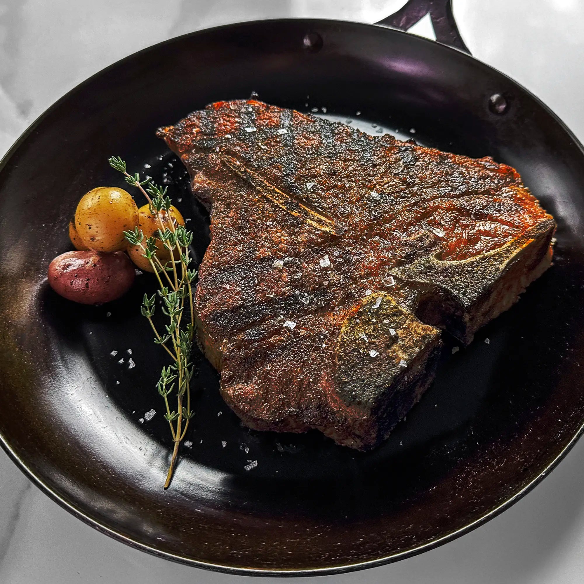 Seared bone-in steak in a dark pan with coarse salt, accompanied by red and yellow baby potatoes, and a sprig of fresh thyme.
