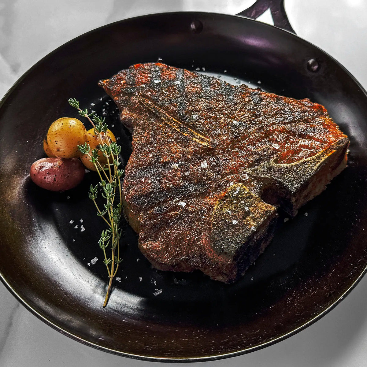 Seared bone-in steak in a dark pan with coarse salt, accompanied by red and yellow baby potatoes, and a sprig of fresh thyme.