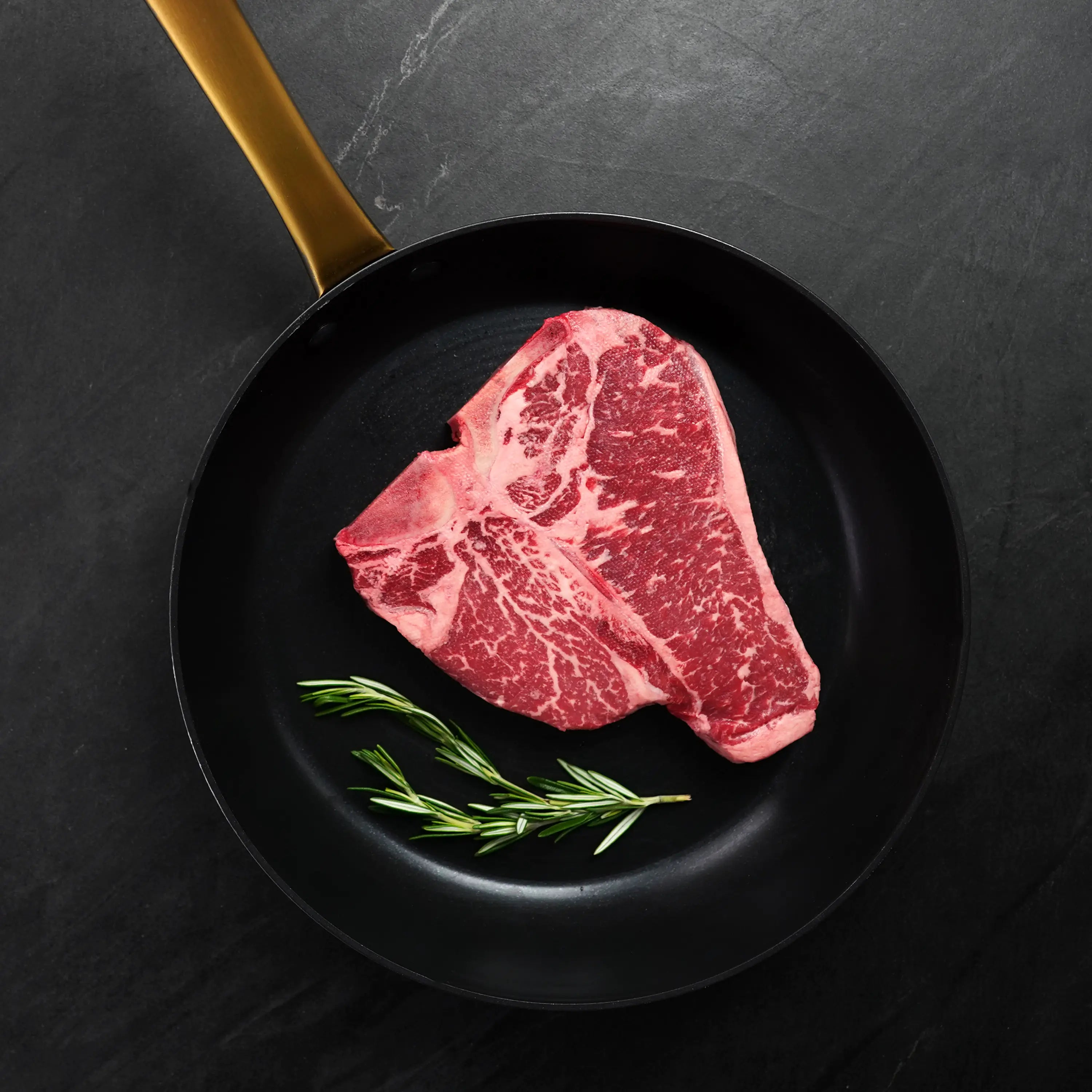 Raw, well-marbled T-bone steak in a black skillet with brass handle, accompanied by fresh rosemary, set on a dark stone background.