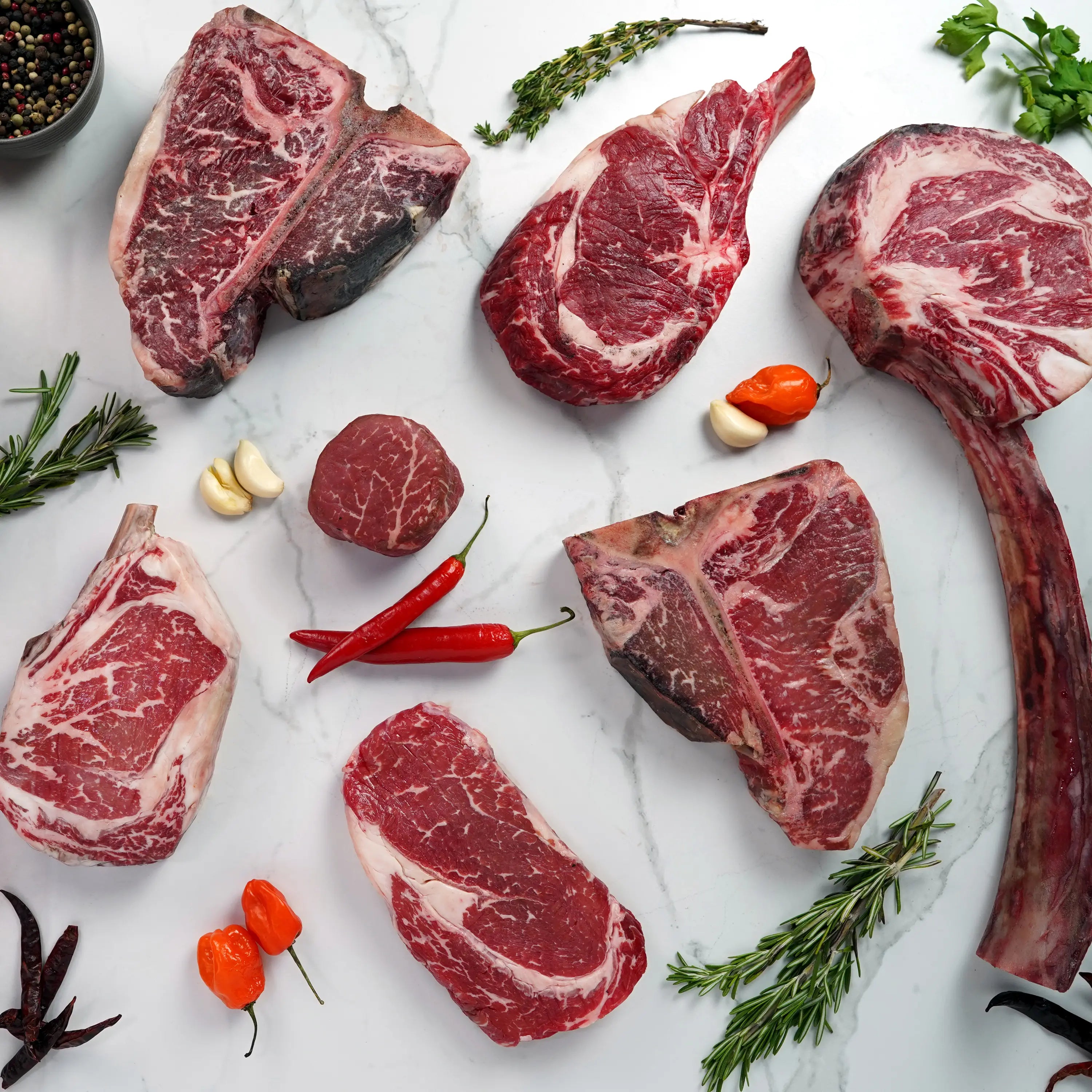 Assorted raw beef steaks and chops on a marble surface, surrounded by rosemary, thyme, mixed peppercorns, garlic cloves, and red chile peppers.