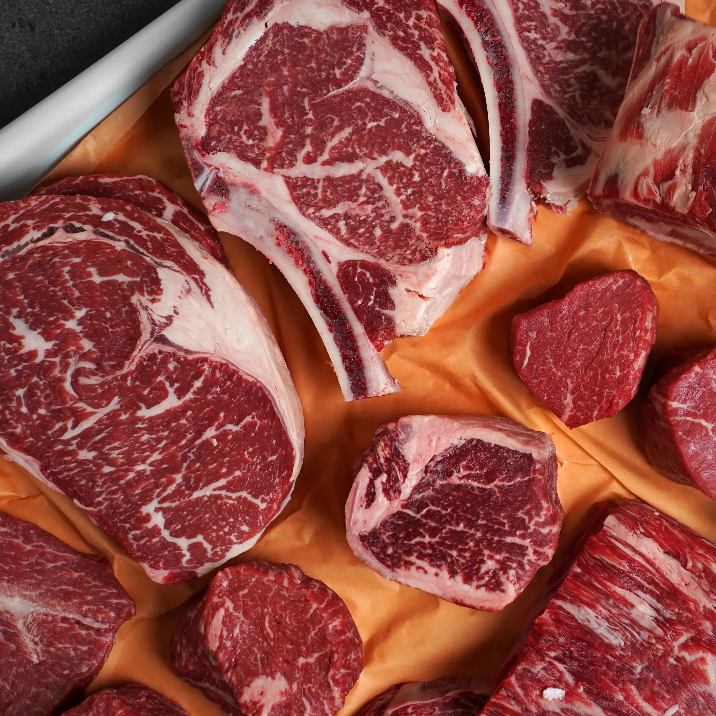 Several cuts of raw, well-marbled beef, including ribeye and other premium steaks, displayed on butcher's paper.
