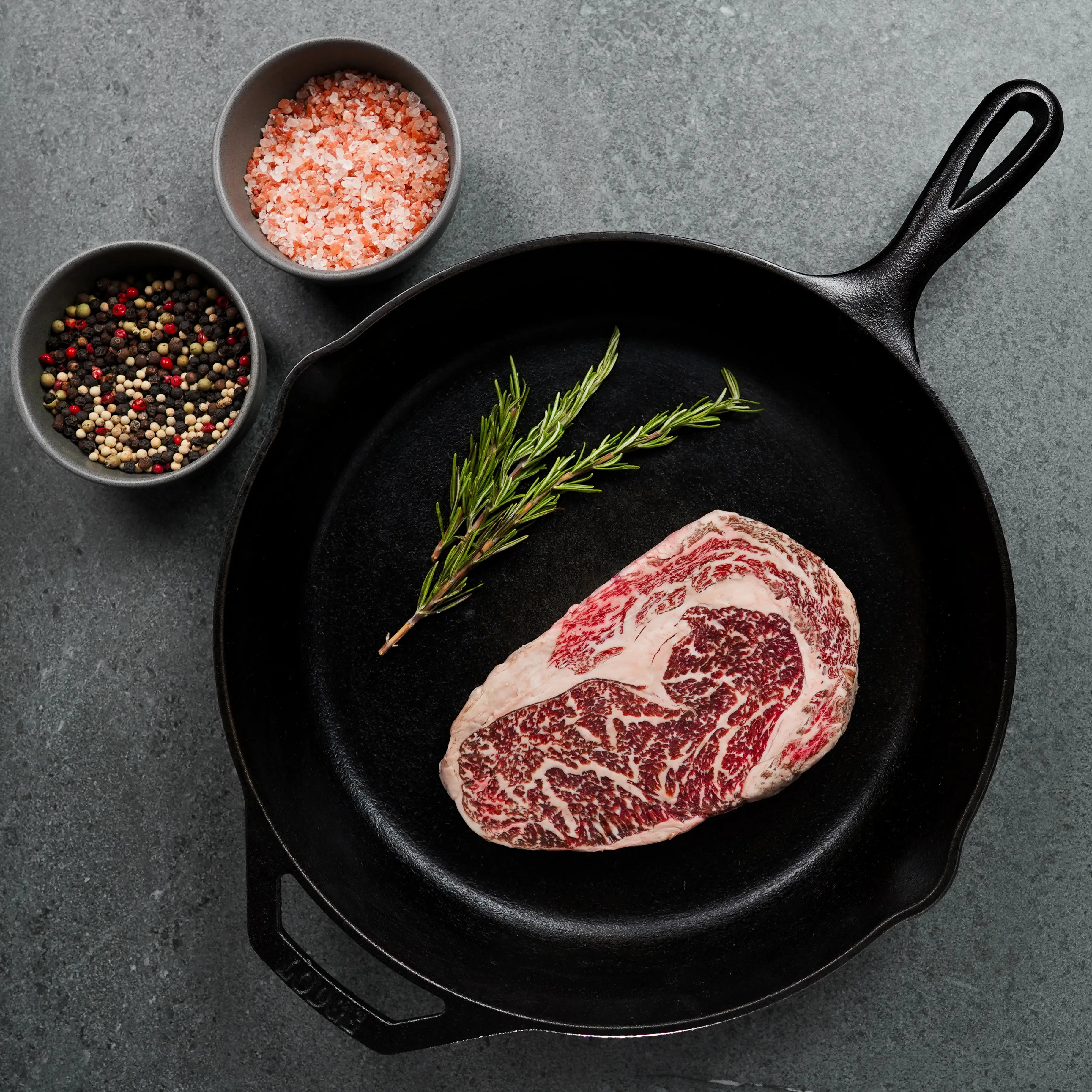 Cast-iron skillet with a raw rib-eye steak and rosemary, accompanied by bowls of pink salt and mixed peppercorns on a stone countertop.