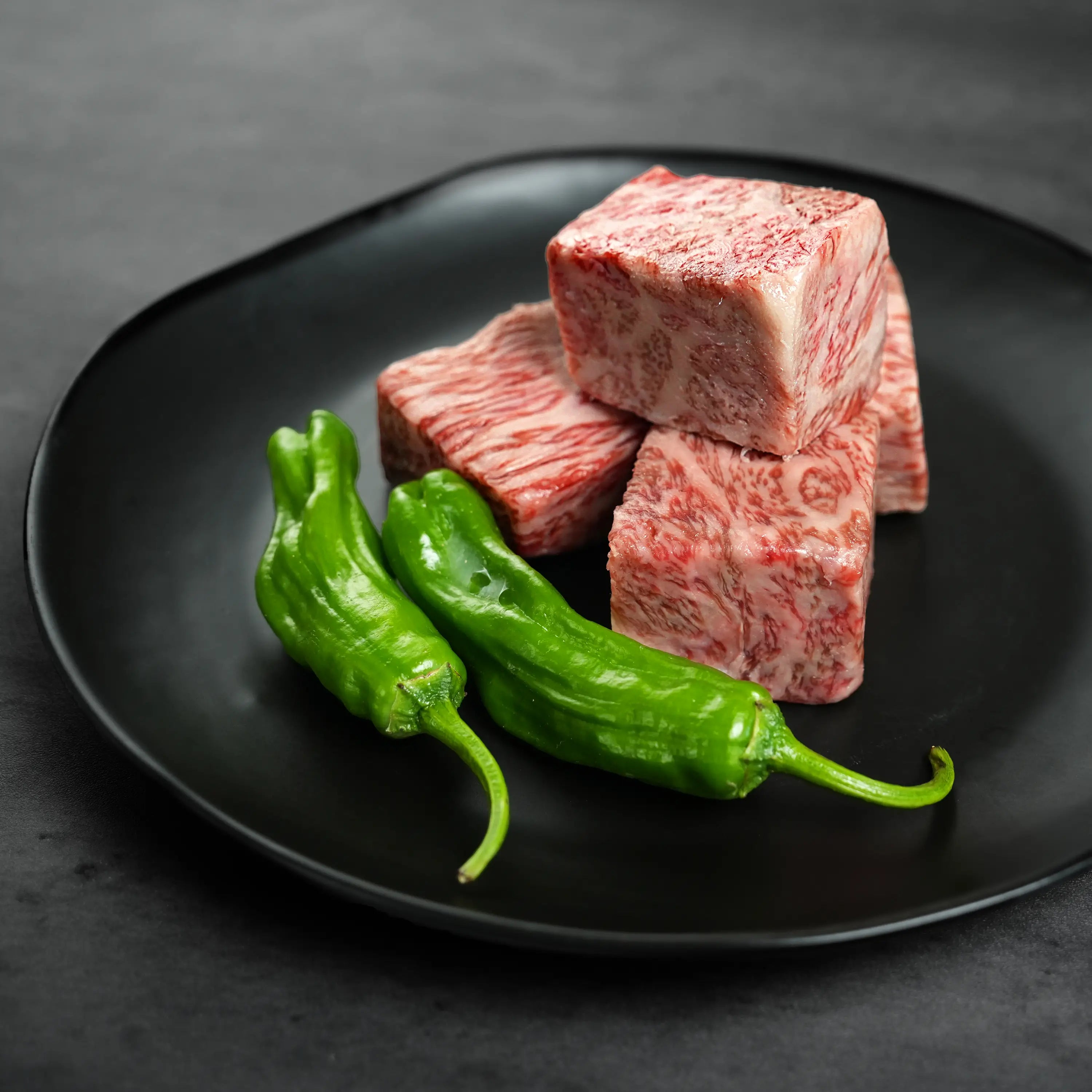 Black plate with marbled raw Wagyu beef cubes and glossy green shishito peppers, highlighting premium meat quality and fresh produce.