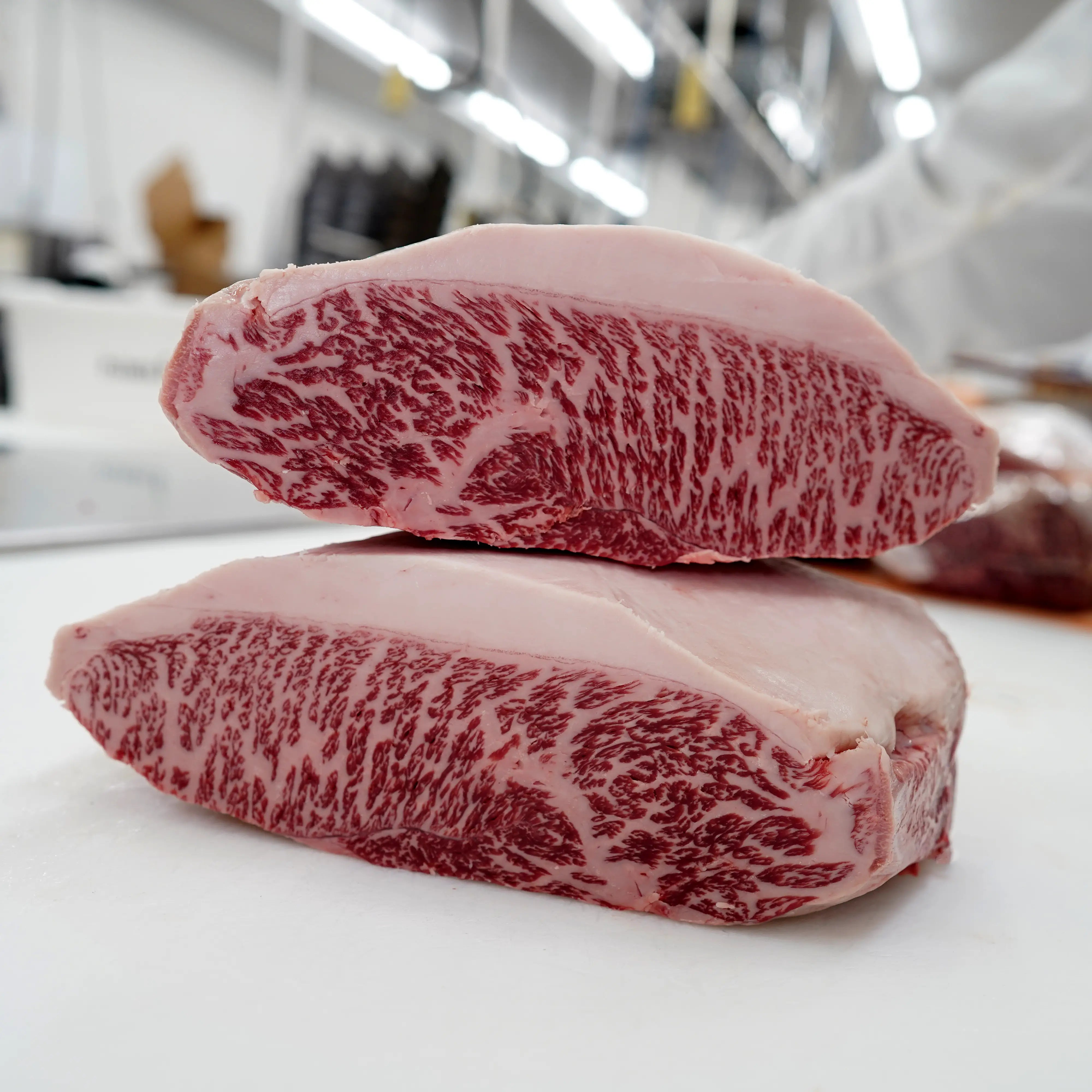 Two raw Wagyu beef steaks with heavy marbling and creamy fat cap, displayed on a white cutting surface in a butchery setting.