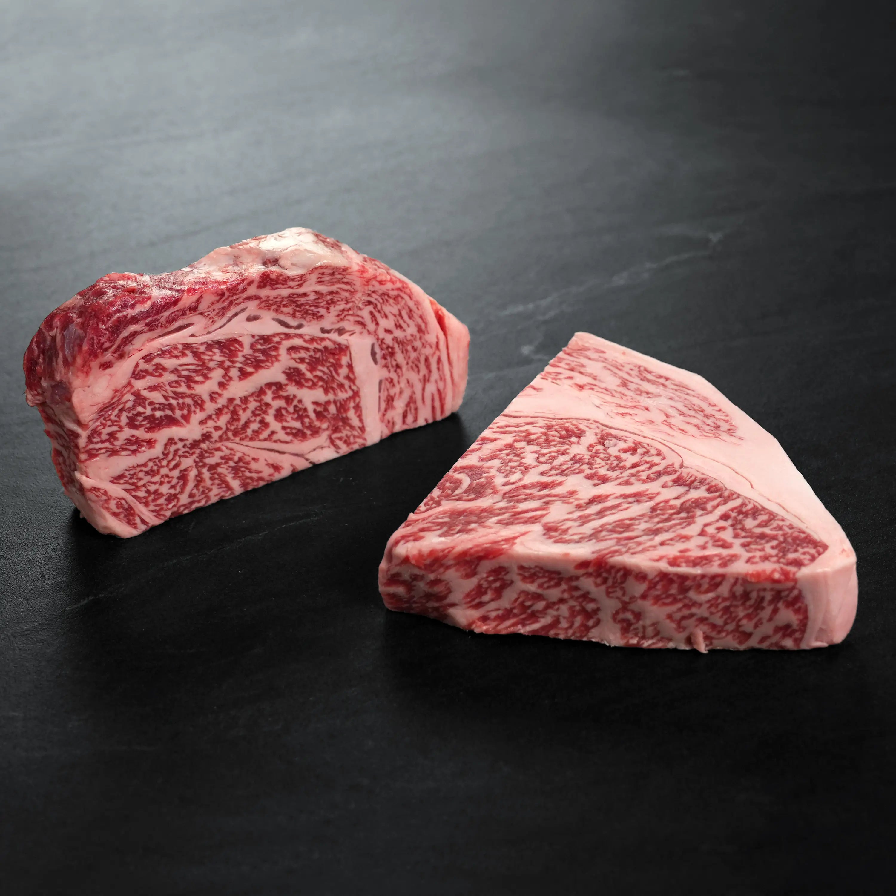 Two thick cuts of raw, highly marbled beef, showcasing intricate marbling patterns, rest on a dark surface, highlighting their premium quality.