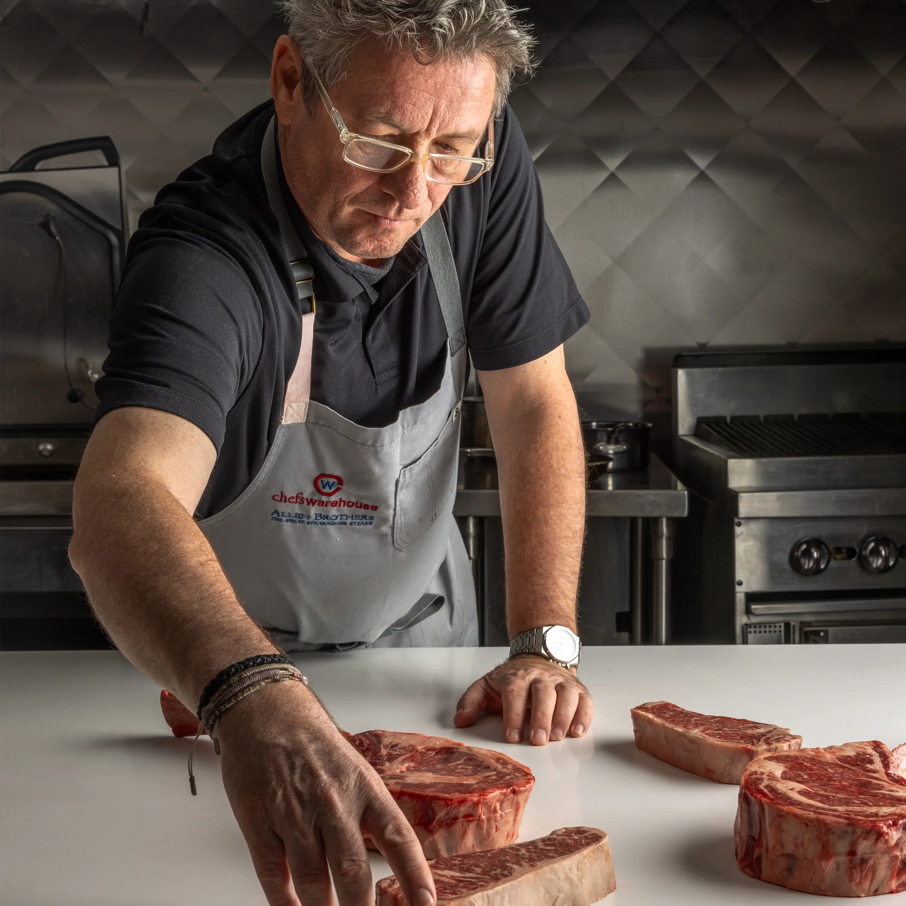 Chef in apron arranging thick cuts of raw steak on a white surface in a professional stainless-steel kitchen with grill equipment visible.
