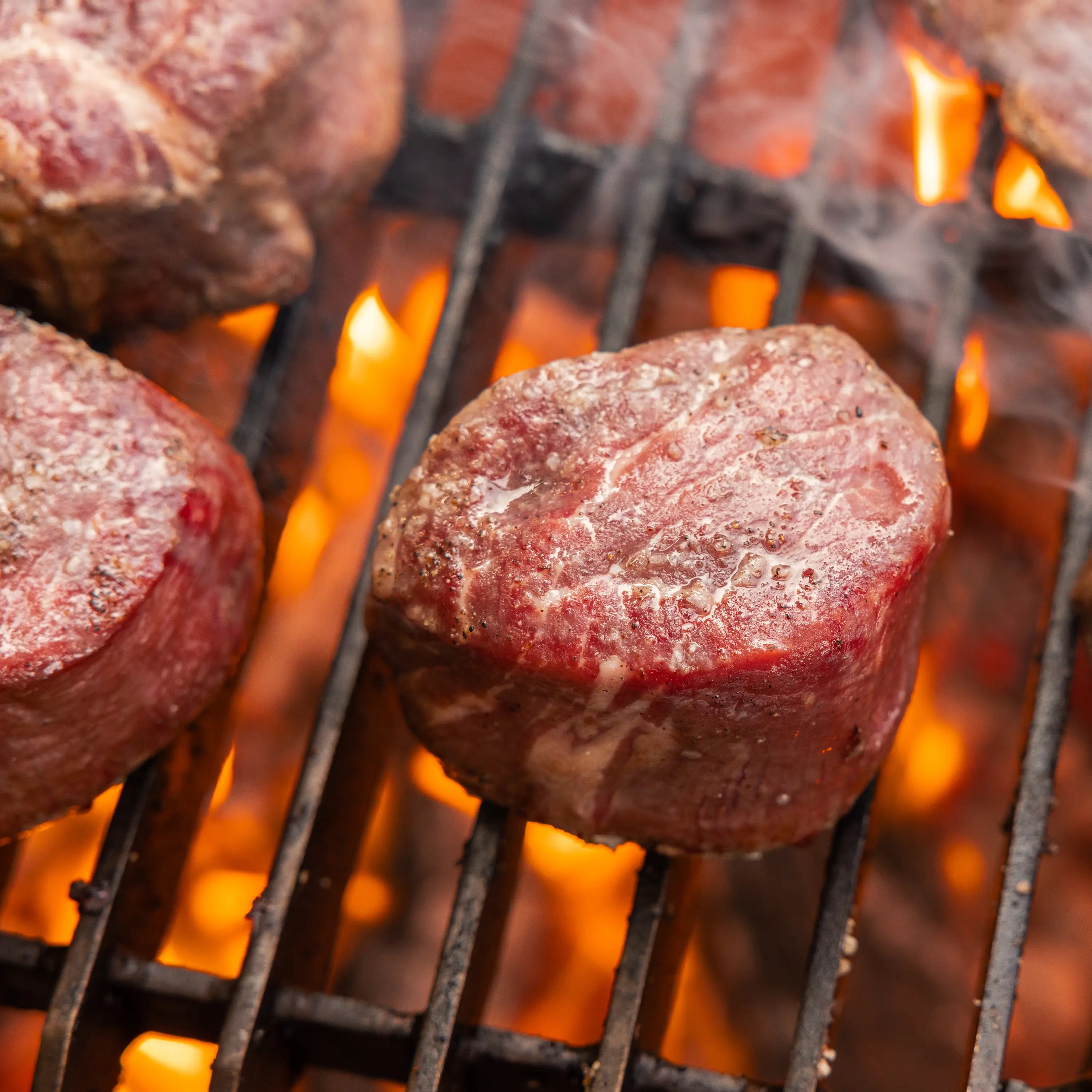 Thick beef steak medallions sear on a hot grill, flames rising through the grates, forming a crispy crust on the meat.