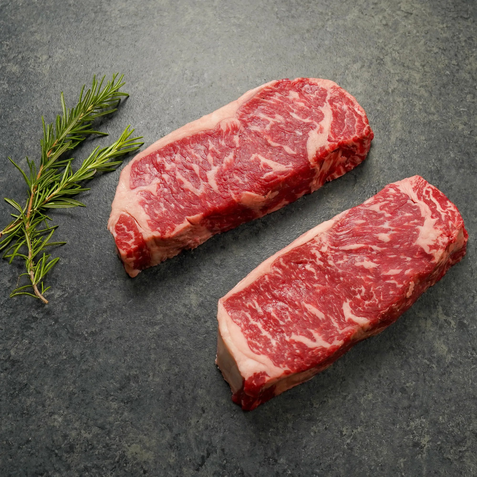 Two thick, well-marbled raw beef steaks on a dark surface with a sprig of fresh rosemary beside them.