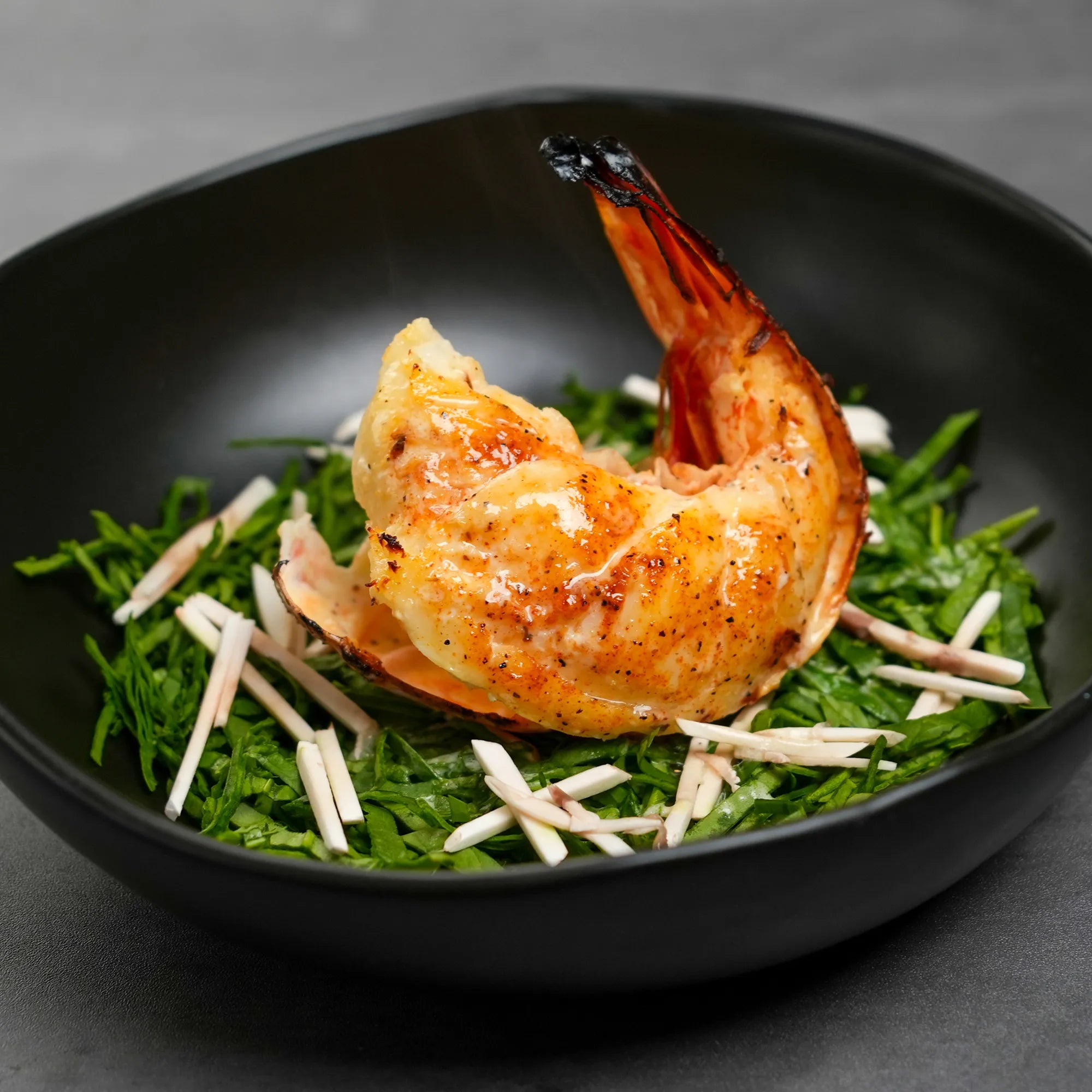 Butterflied grilled prawn in shell atop chopped samphire and white vegetable strips, presented in a shallow matte-black bowl with light seasoning.