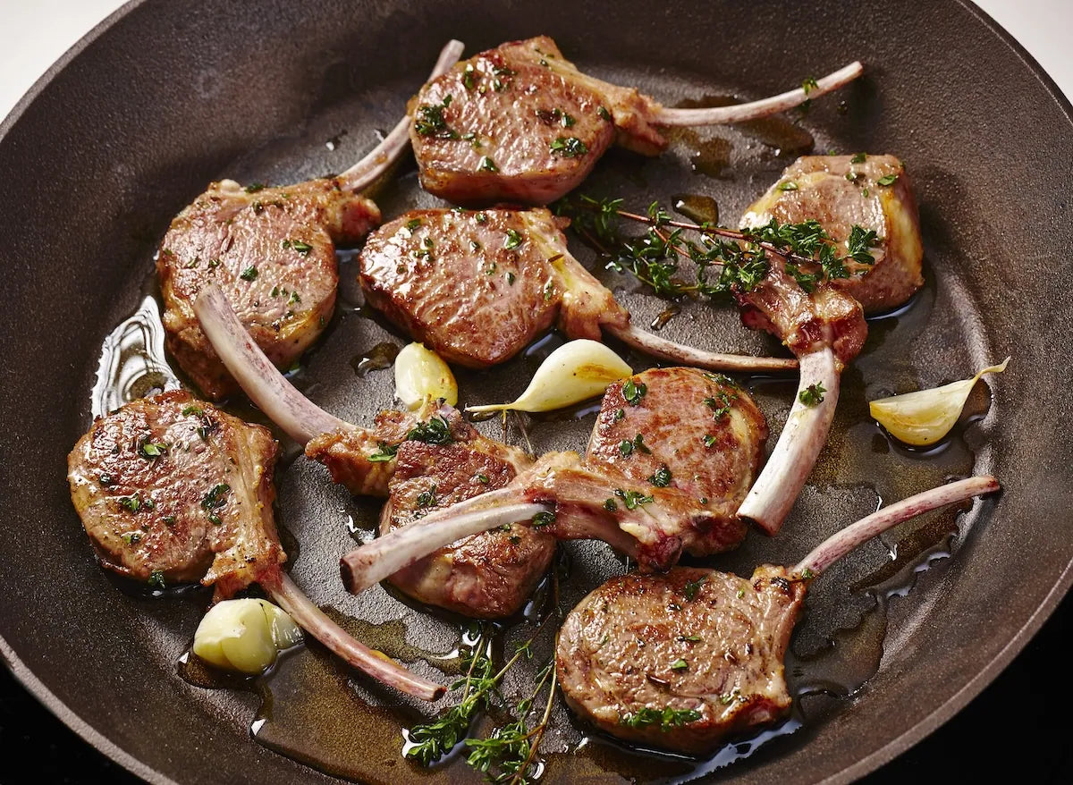 Pan-seared lamb chops with bone-in bones, cooked in a dark cast-iron skillet with garlic cloves, fresh thyme, and oil.