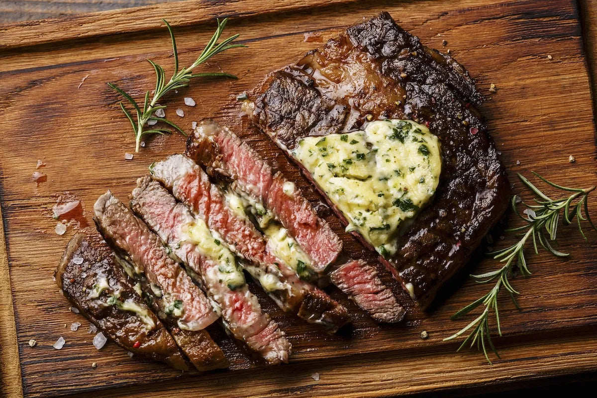 Sliced medium-rare steak topped with a dollop of herb butter on a rustic wooden cutting board, garnished with rosemary sprigs and coarse salt.