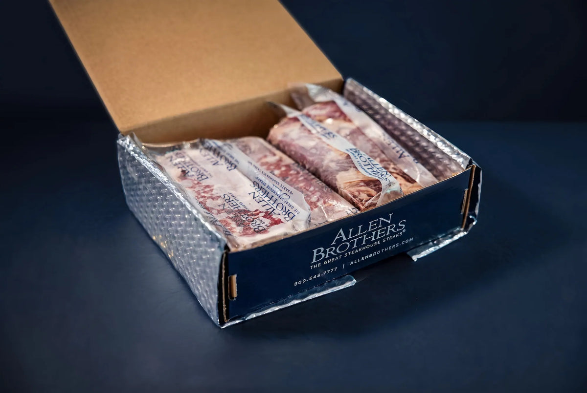 An open cardboard box containing several packaged sliced meats, with a dark navy branded outer box visible on a dark blue background. The brand name Allen Brothers is clearly printed on the front.