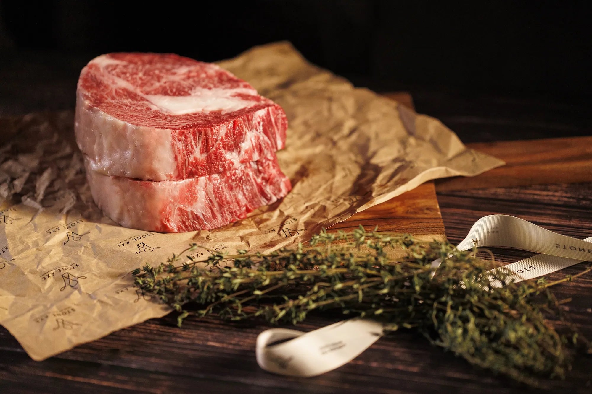 Two thick slices of marbled raw beef resting on brown paper over a wooden surface, with a sprig of fresh herbs and a white ribbon in the foreground.