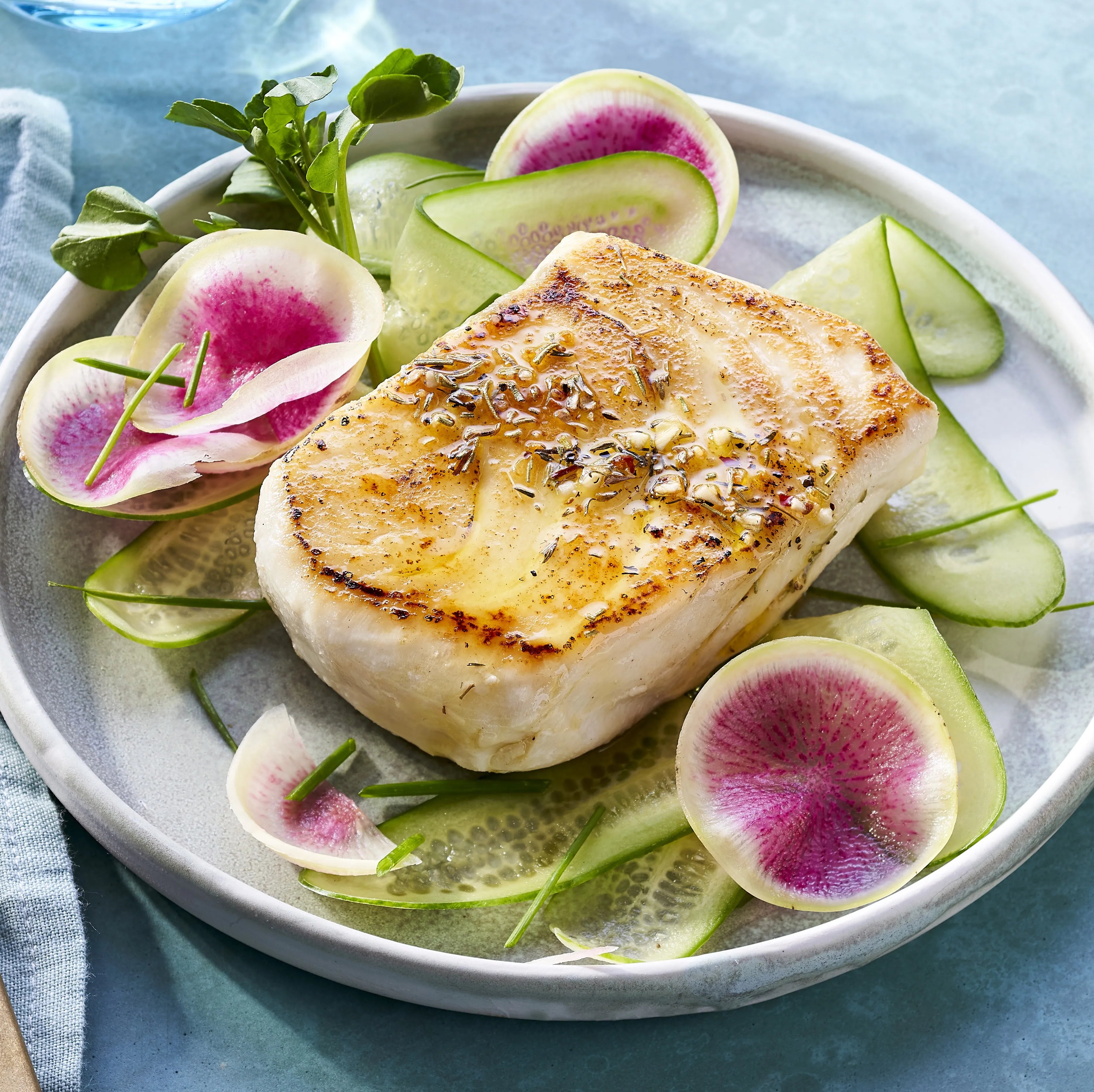 Seared fish fillet on a bed of sliced cucumbers and watermelon radishes on a white plate.