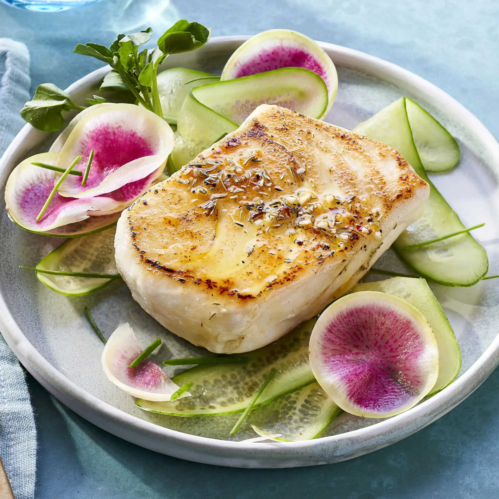 Seared fish fillet on a bed of sliced cucumbers and watermelon radishes on a white plate.