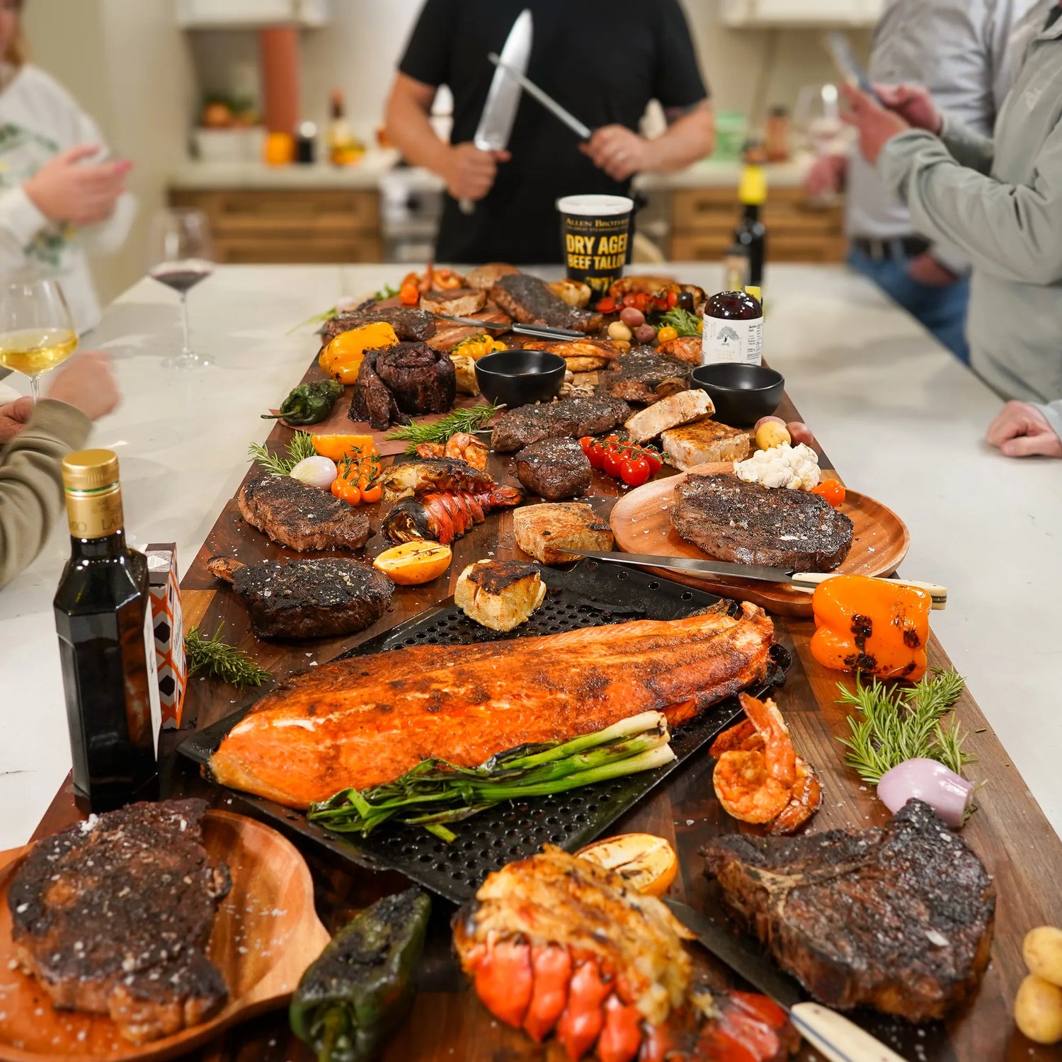 A wooden board with grilled salmon, dry-aged steaks, lobster tails, shrimp, roasted vegetables, and herbs, surrounded by condiments, wine glasses, and people preparing to serve.
