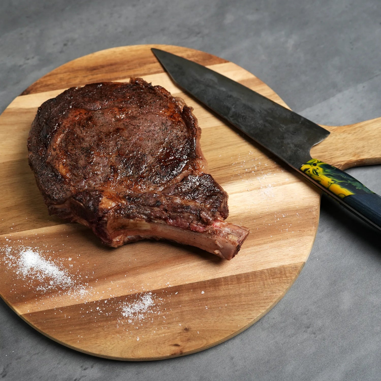cooked steak on wood board with salt and knife next to it on light surface