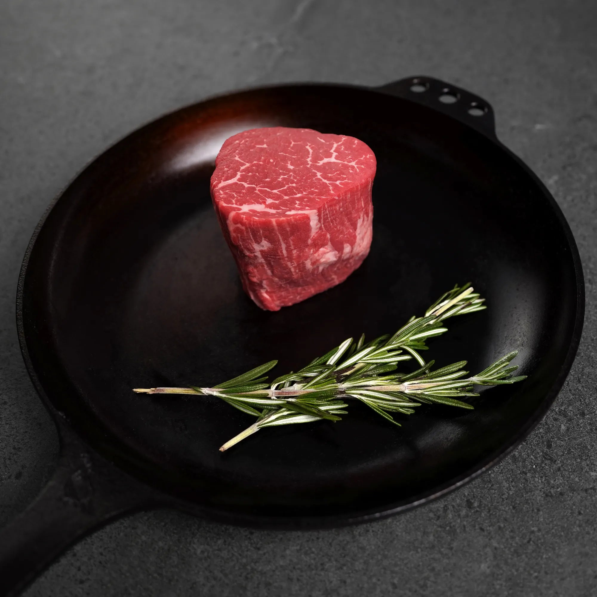 raw steak on cast iron pan with green garnish on dark surface