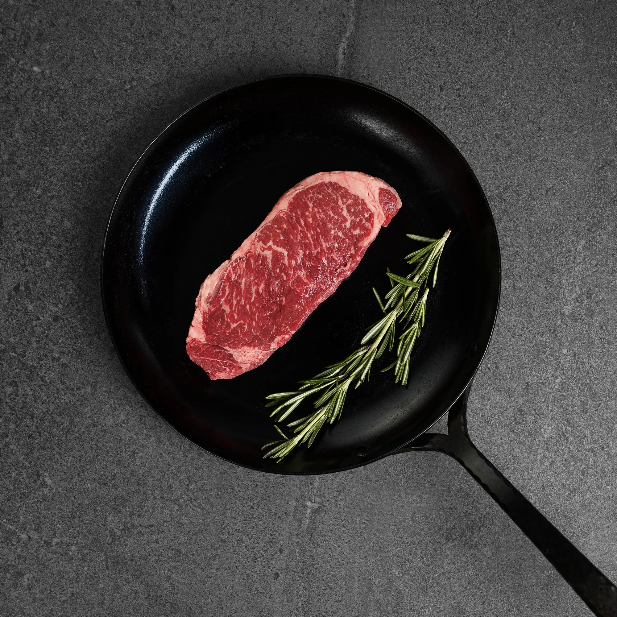 raw strip steak on cast iron pan with rosemary garnish