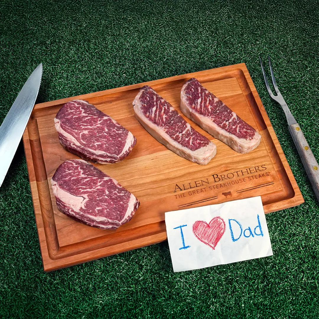Wooden cutting board with Allen Brothers branding featuring two rib-eye and two strip steaks, carving knife, meat fork, and a crayon-drawn “I ❤️ Dad” note.