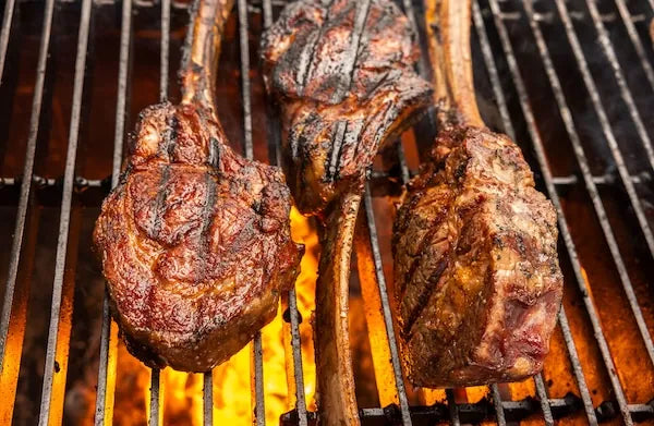 Three large tomahawk ribeye steaks sear over hot coals on a barbecue grill, each displaying char lines from the grates.