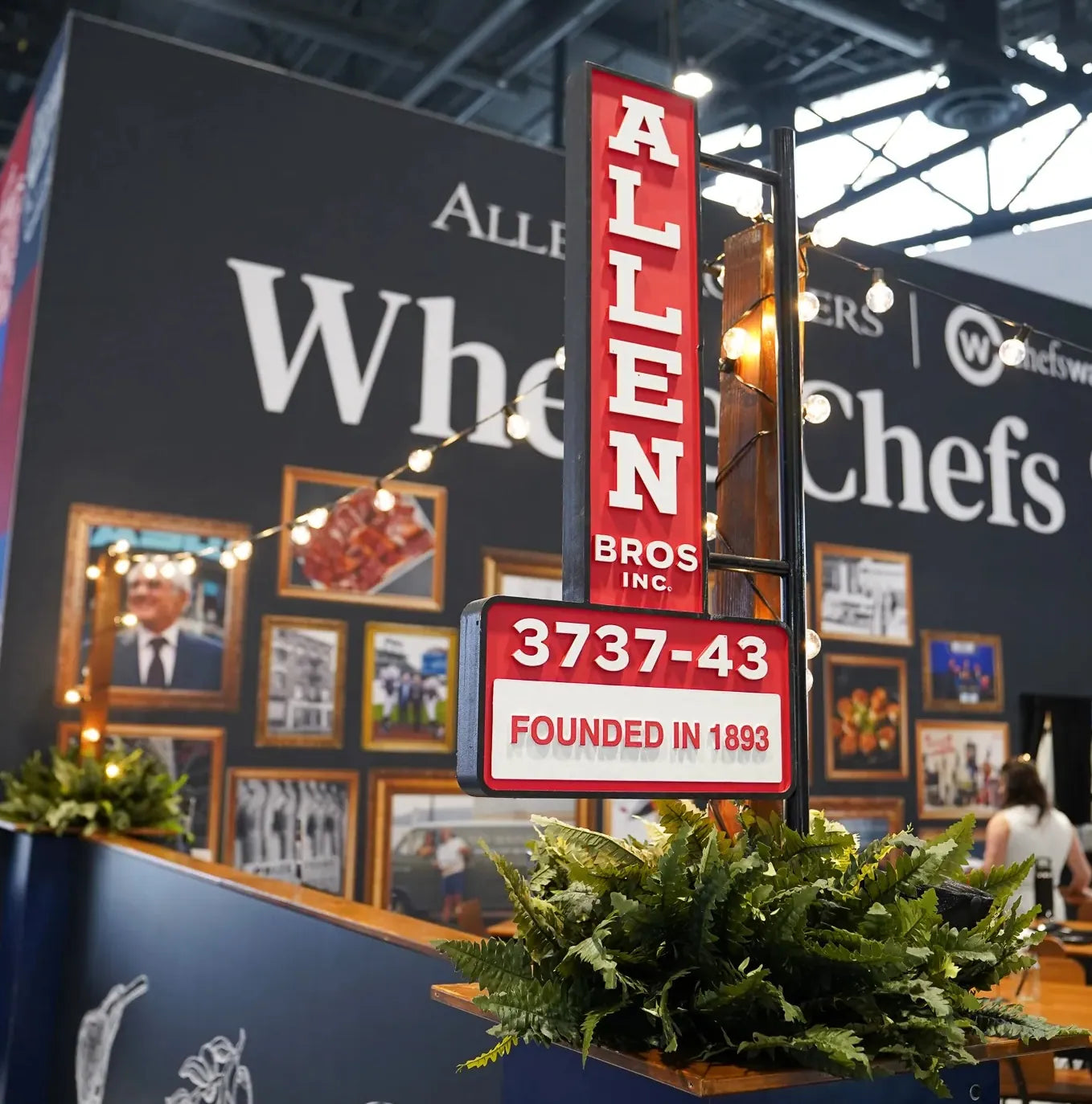 Trade-show booth with a tall red sign for Allen Bros Inc., founded 1893, surrounded by string lights and green ferns. Framed photos in background.