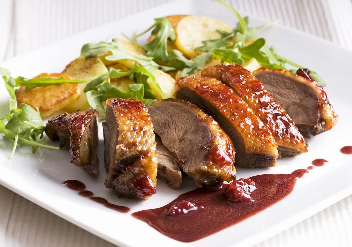 Sliced roasted duck with a glossy browned skin and dark red sauce on a white plate, served with green salad and potato wedges.