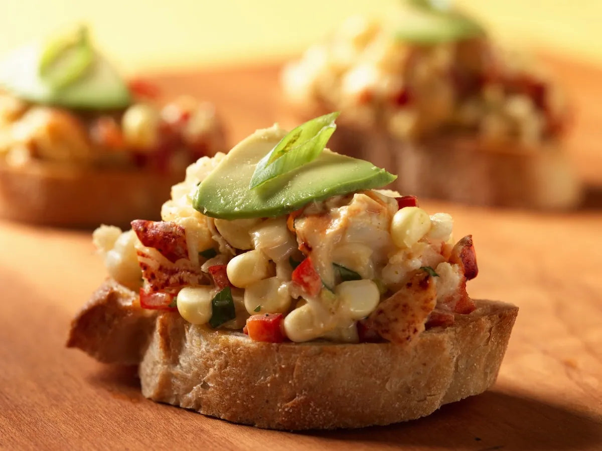 A close-up shot of a piece of toasted bread topped with a creamy corn and bacon salad, garnished with fresh avocado slices, set on a wooden surface with other blurred servings in the background.