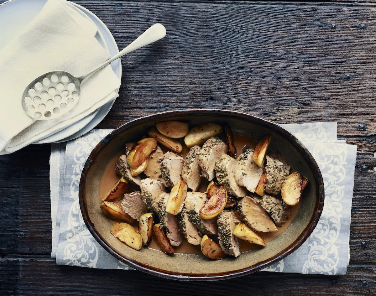 An oval ceramic baking dish filled with sliced roasted meat and potato wedges, set on a patterned napkin over a dark wooden table. A folded white napkin with a metal spoon rests in the upper left.