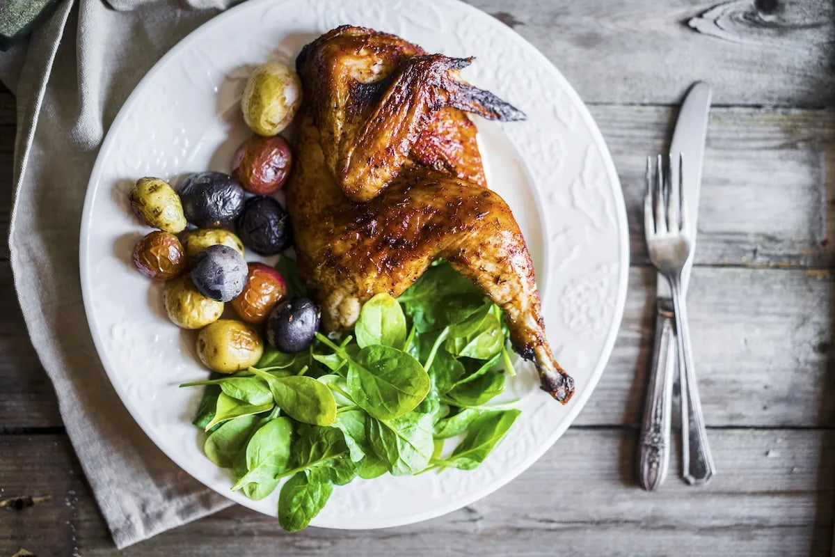 A roasted whole chicken served on a white plate with green salad greens, roasted small potatoes, and a fork and knife on a rustic wooden table.