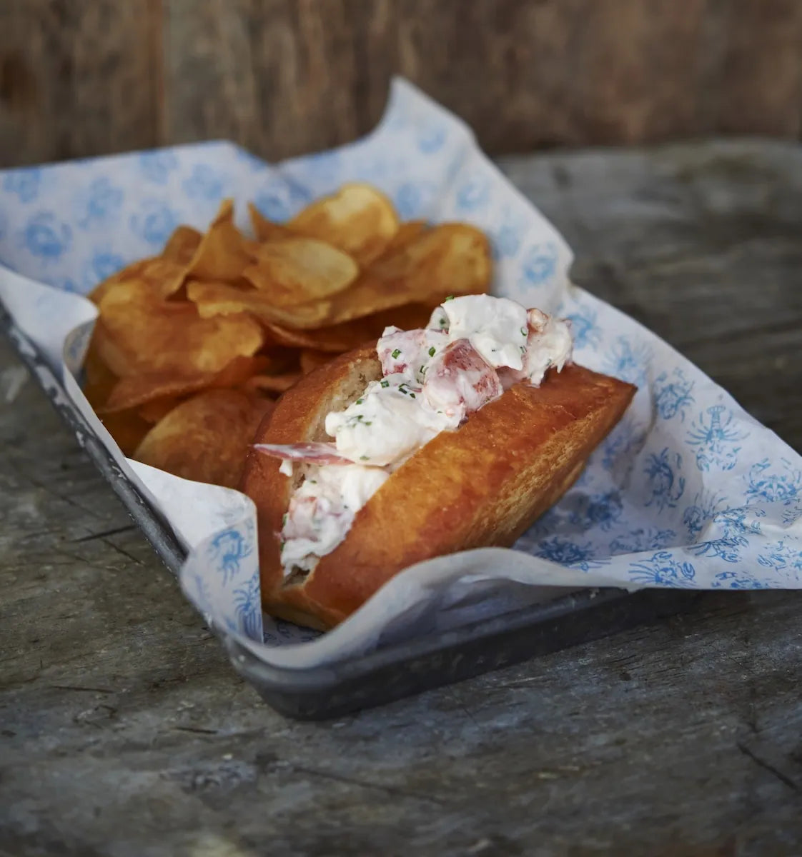 A lobster roll sandwich in a bun with a creamy white lobster mixture and red onion, served with potato chips in a clear plastic tray lined with blue-and-white patterned paper on a rustic wooden surface.