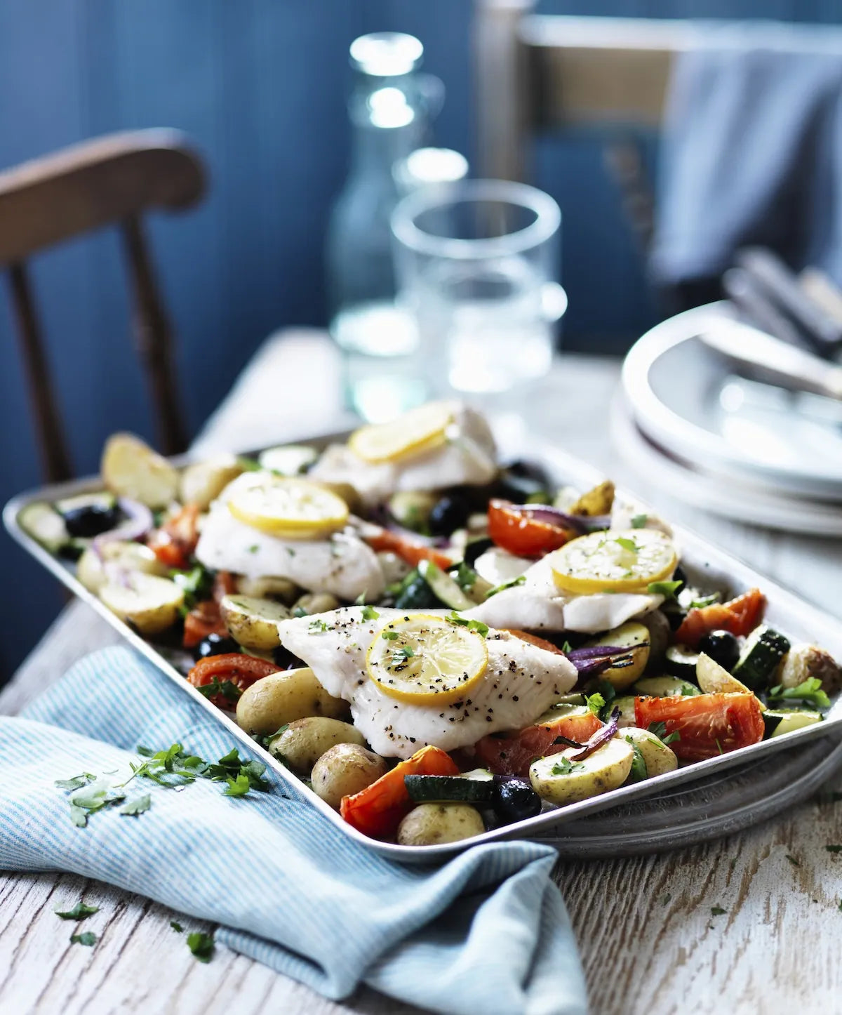 A roasted fish and vegetable dish served on a rectangular metal tray, set on a rustic table with a blue napkin, glass bottle, and plates in the background.