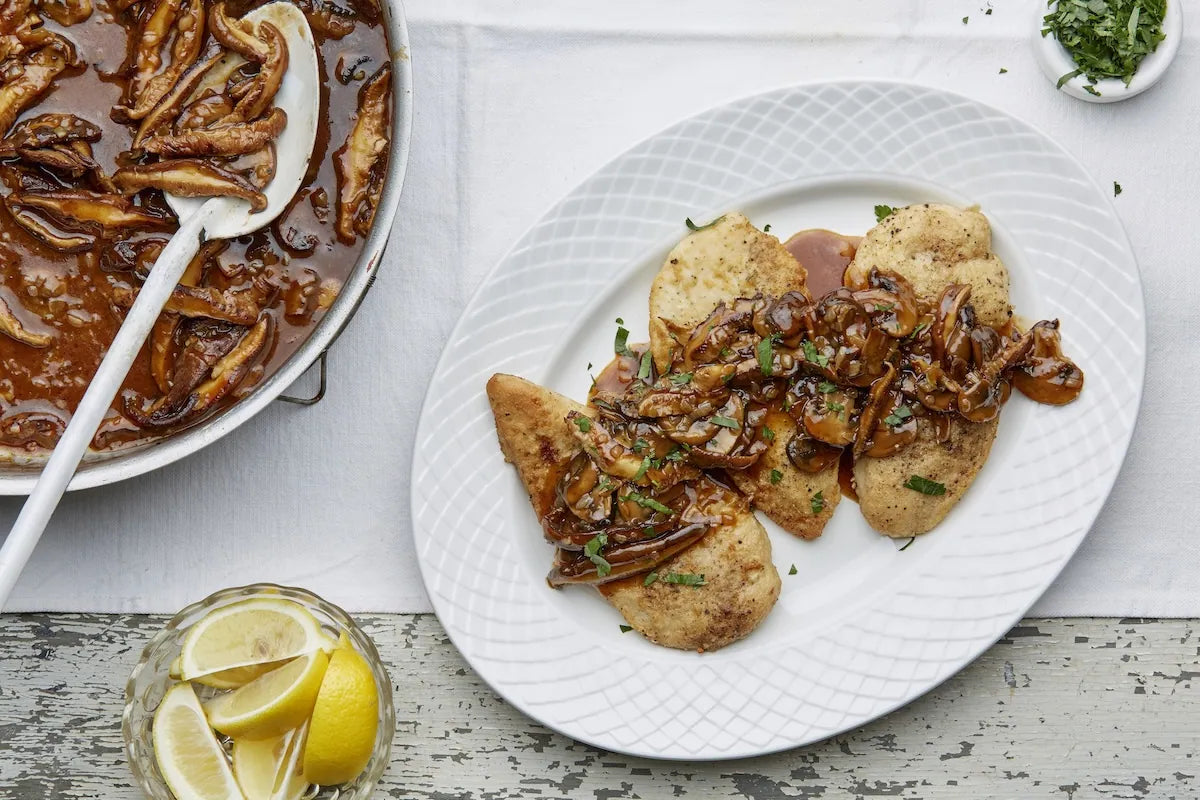 A plated dish of chicken cutlets topped with caramelized onions and a brown sauce, served on a white patterned plate. A large pan of mushrooms in a brown sauce with a white spoon is visible on the left, along with lemon slices in a glass and a small dish of herbs on a white tablecloth.