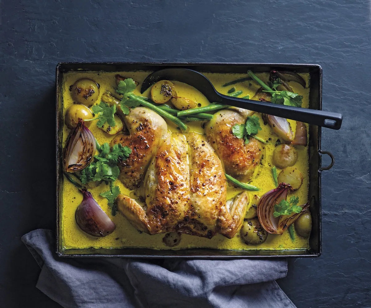 A roasted whole chicken in a yellow-orange sauce with potatoes, red onions, green beans, and herbs, served in a dark rectangular baking pan on a dark slate surface with a gray cloth underneath.