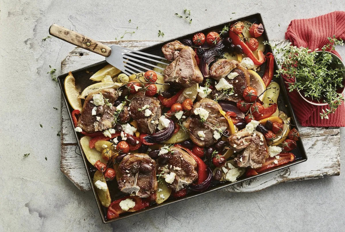 A roasted meat and vegetable dish served in a rectangular baking pan with a spatula, placed on a rustic board over a light stone surface. A small bowl of fresh herbs and a red cloth napkin sit to the right.