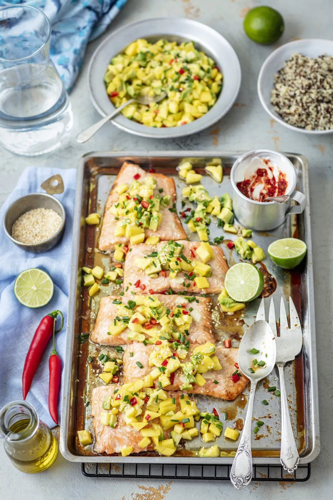 Pan-seared salmon fillets topped with a fresh mango salsa, arranged on a metal baking sheet with forks, spoons, limes, chili peppers, and bowls of ingredients around it on a light gray surface.