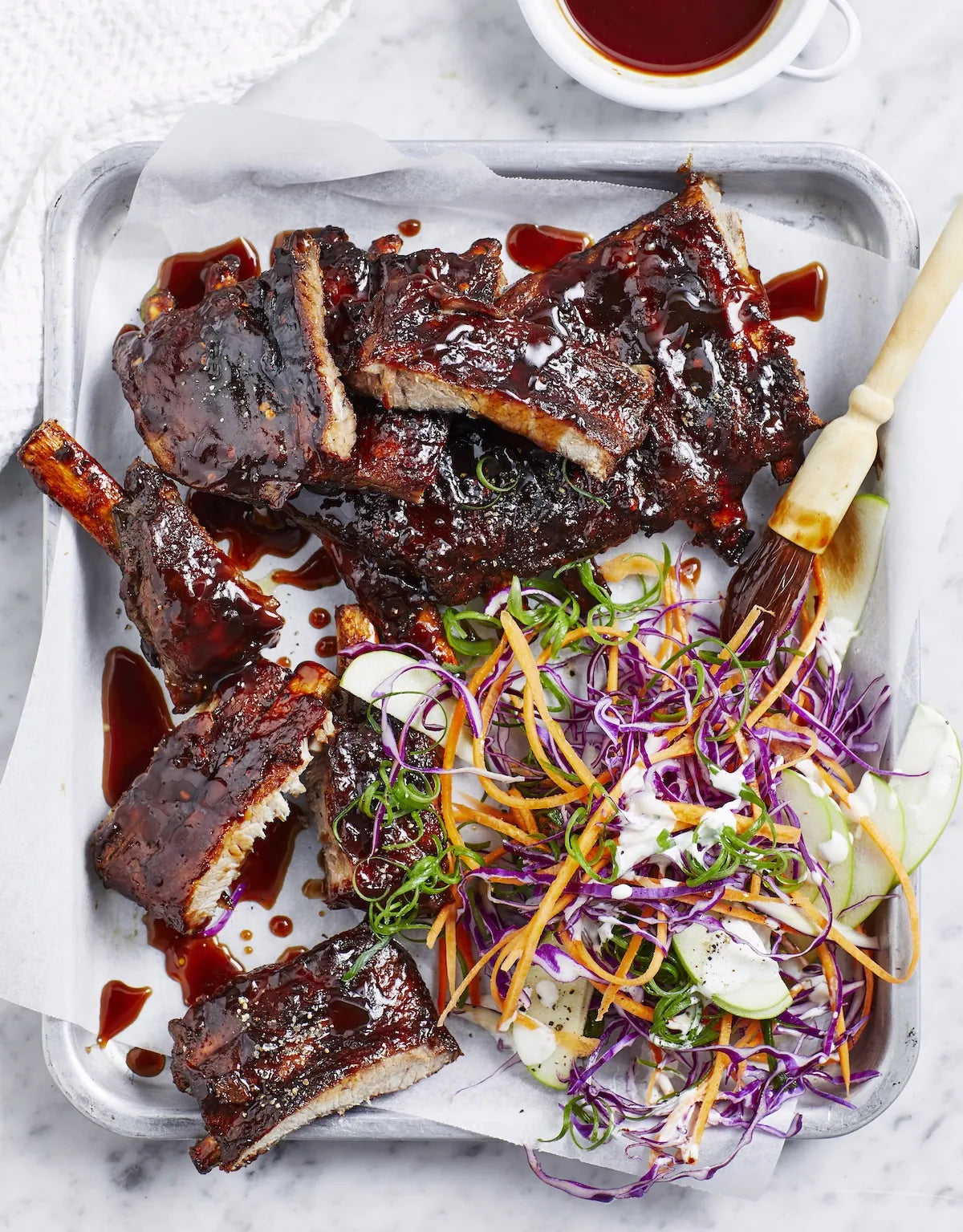 A top-down view of glazed barbecue ribs served on a metal tray with a side salad of shredded purple cabbage, carrots, and cucumber, with a small cup of sauce at the top.