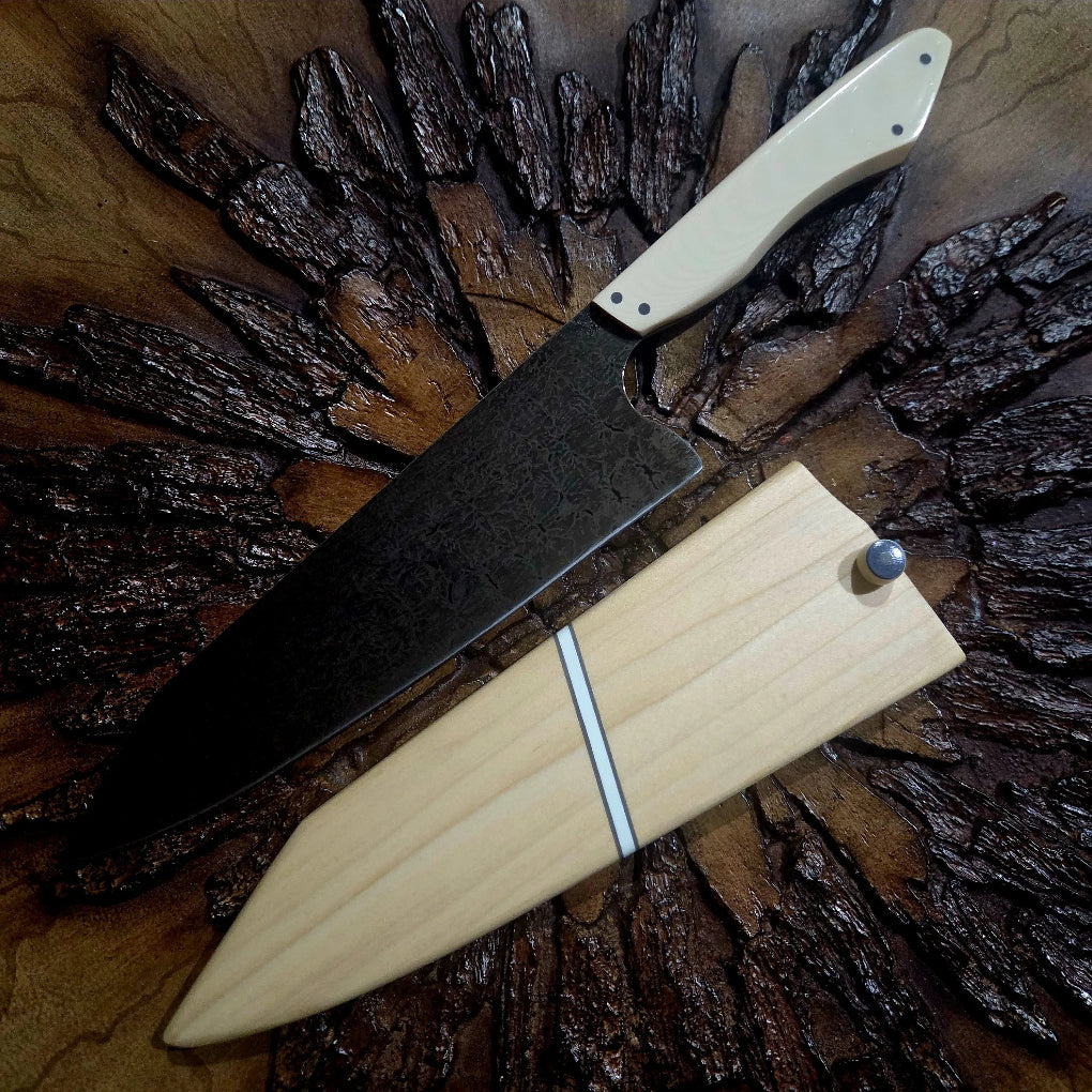 TH Blades Handcrafted 8 Chef Knife with hammered high carbon steel blade and light handle, resting on a textured wooden surface with matching wooden sheath.