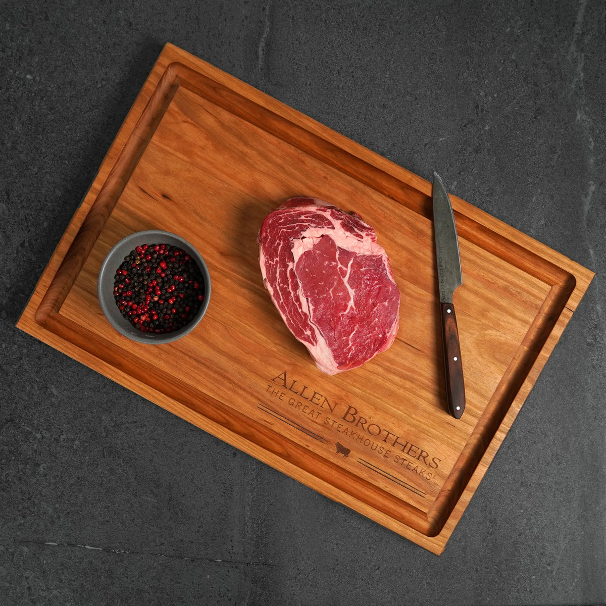 Wooden cutting board with a raw steak, small bowl of spices, and knife on a dark surface.