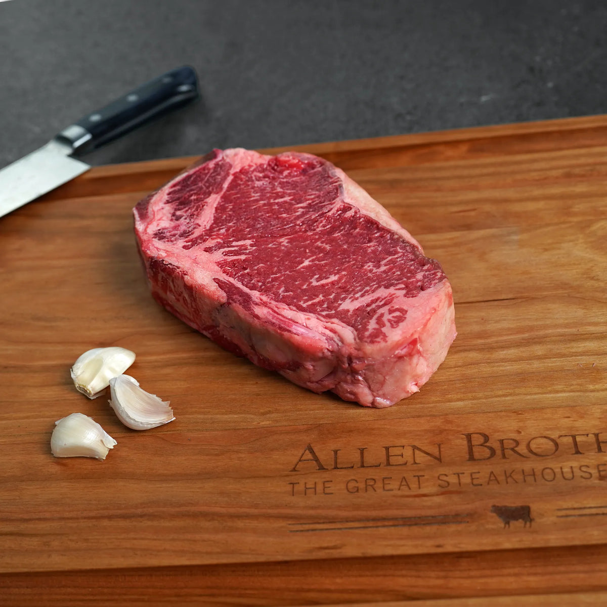 Raw steak on a wooden cutting board with garlic and a knife, branded 'Allen Brothers The Great Steakhouse'.