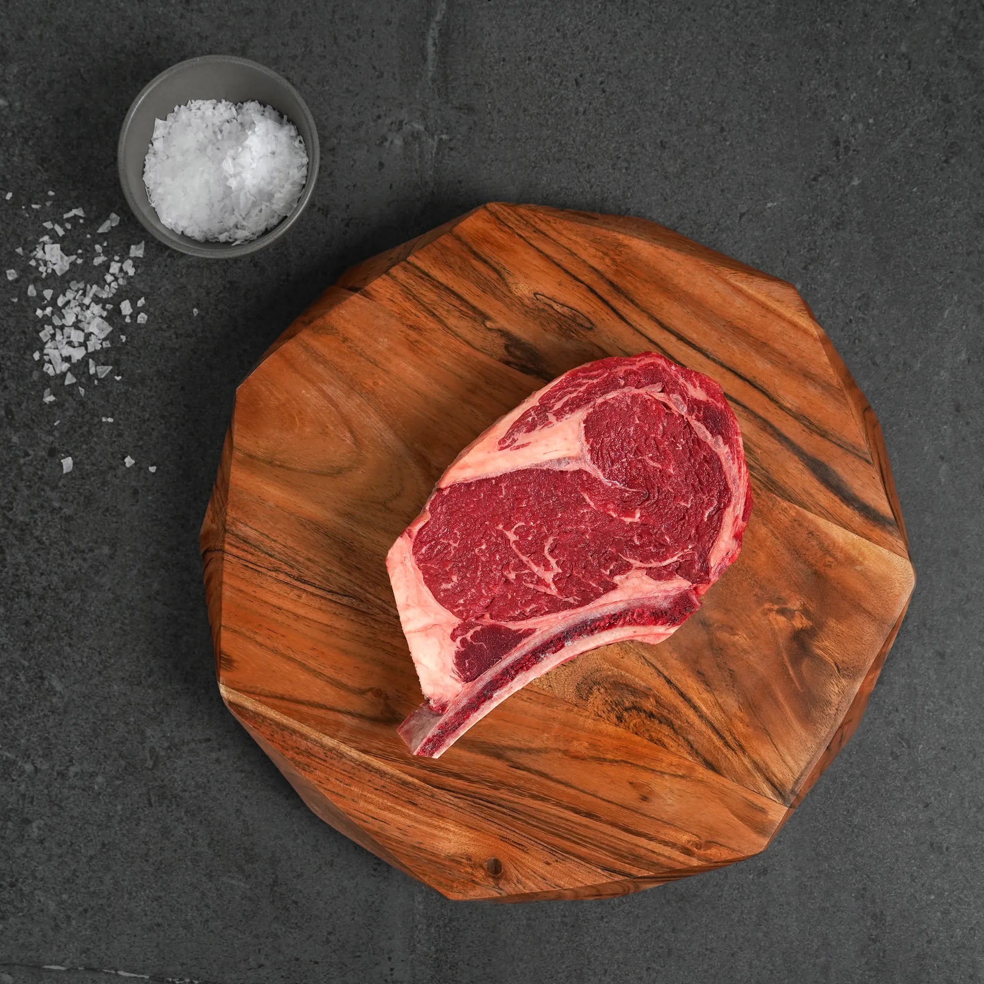 Raw ribeye steak on a wooden cutting board with salt, on a dark surface