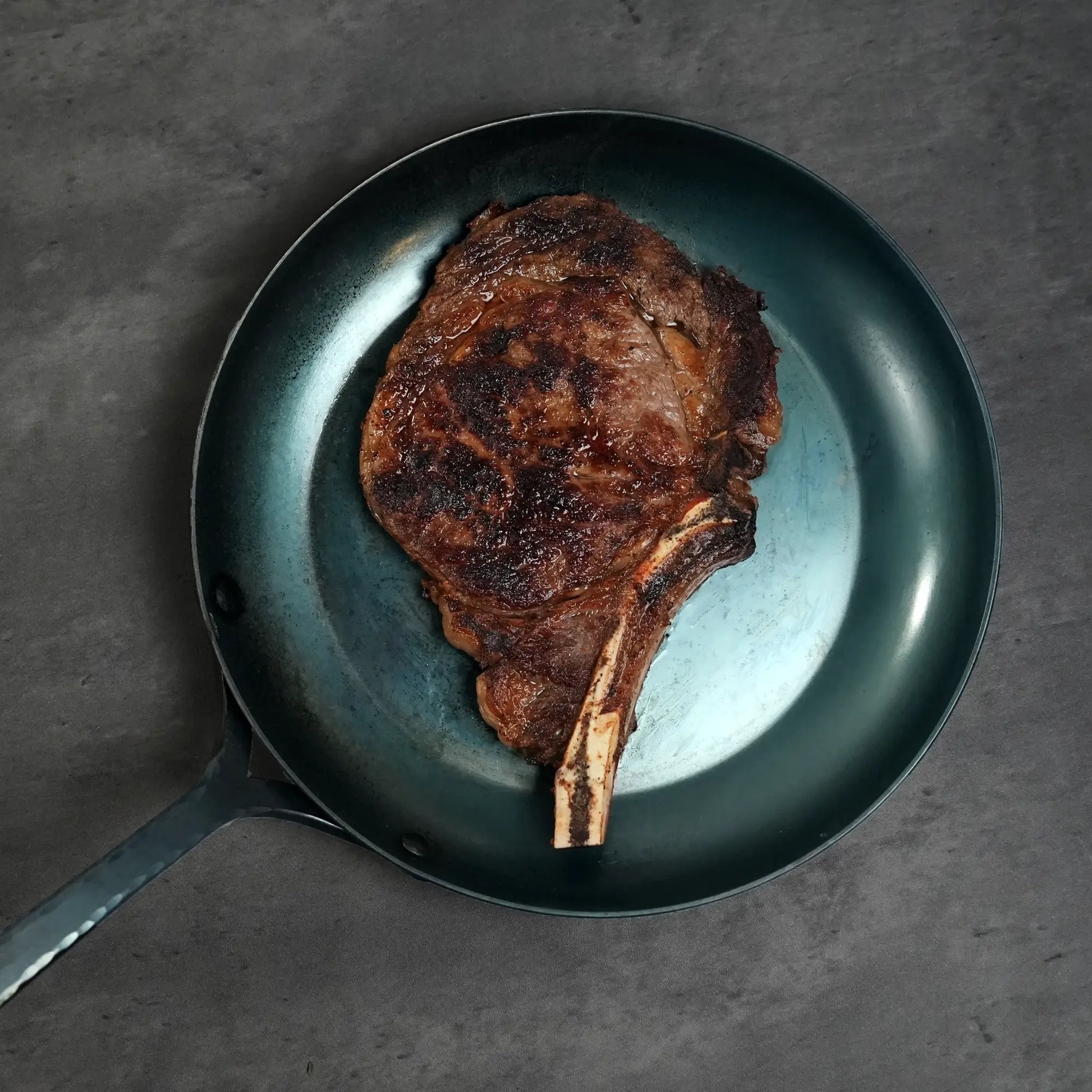 Prime Split-Bone Ribeye Steak seared in a frying pan, showcasing its thick cut for even heat penetration and enhanced flavor, ideal for grilling or broiling.