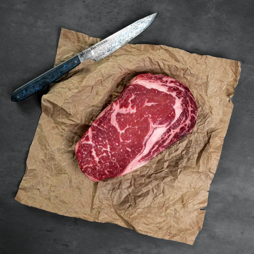 Dry Aged Prime Boneless Ribeye Steak on butcher paper with an Allen Brothers knife, showcasing its marbling, ideal for steak enthusiasts.
