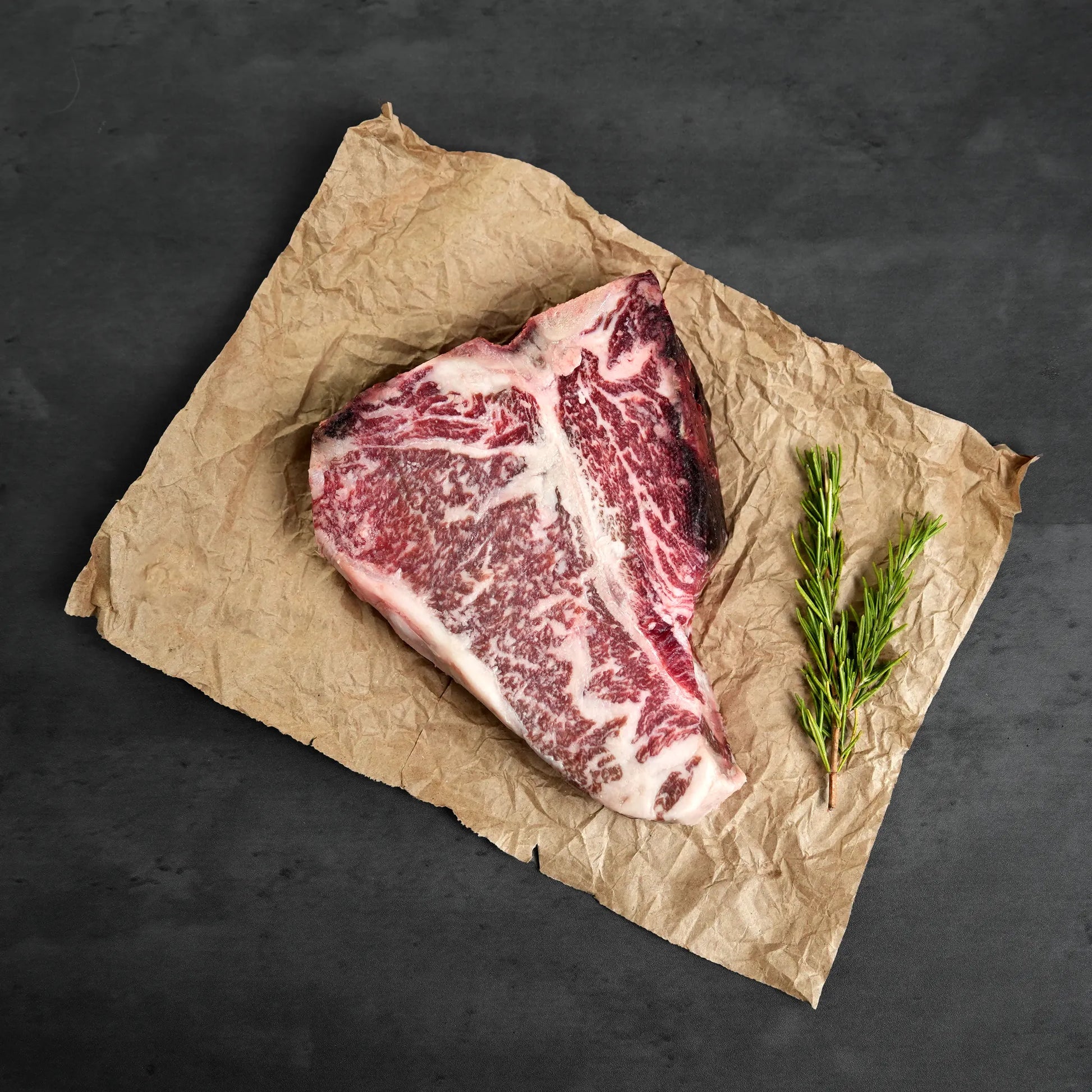 Raw steak on crumpled brown paper with a sprig of rosemary on a dark background