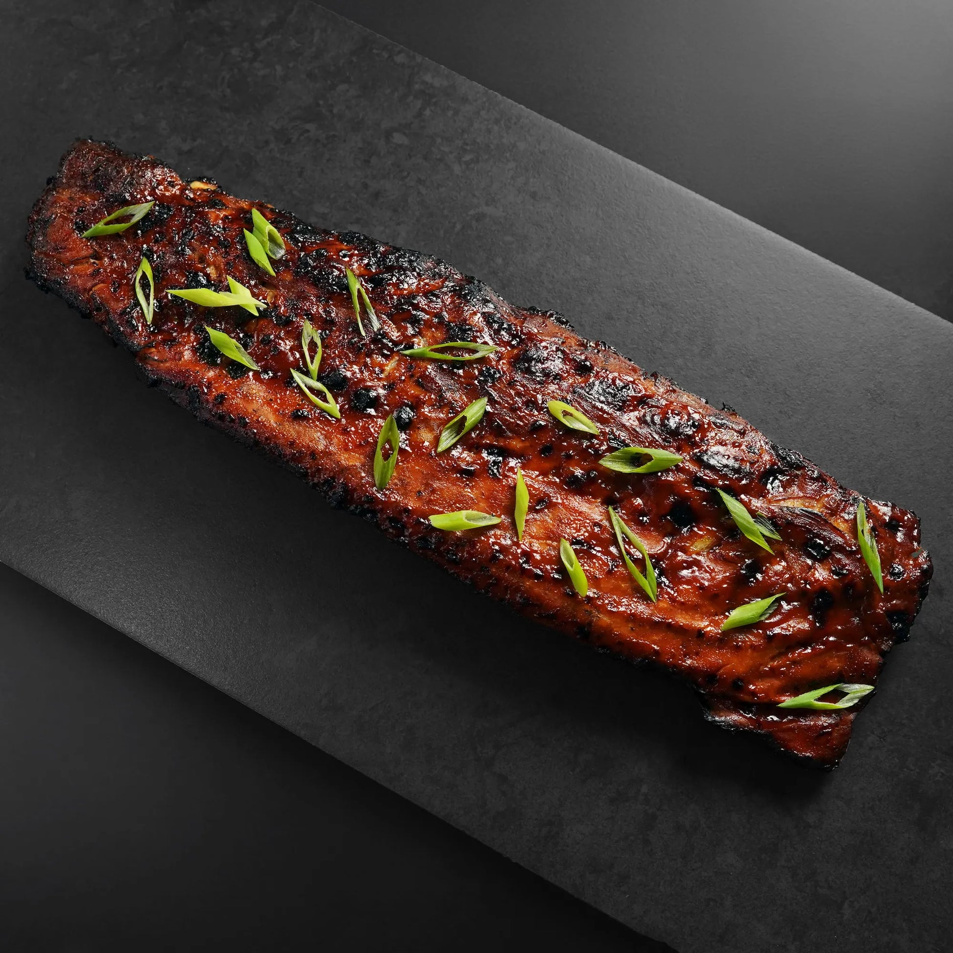 Heritage Duroc Pork Peeled Baby Back Rib, richly glazed and charred, garnished with scallions, presented on a dark platter, showcasing premium barbecue appeal.