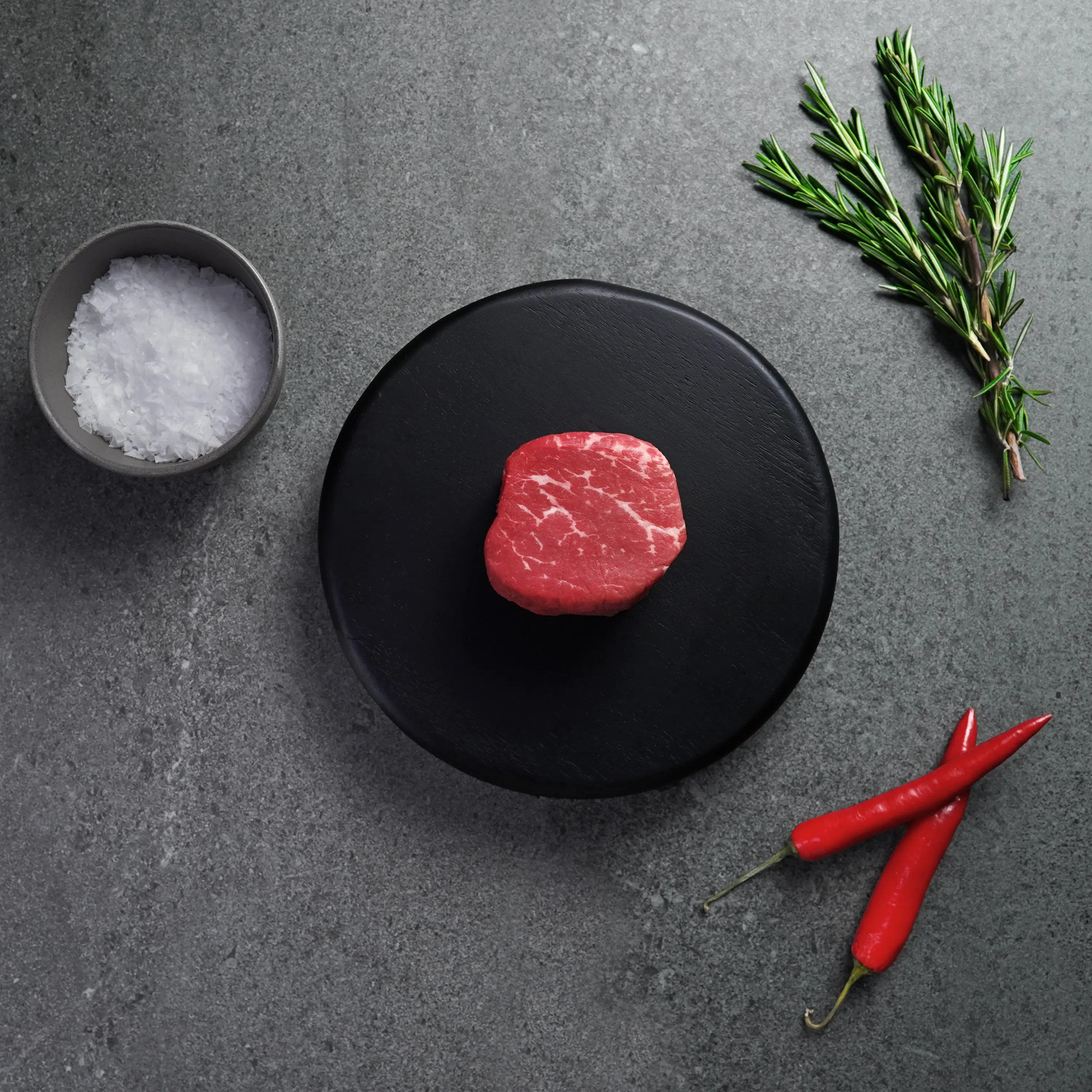 Raw steak on a black plate with salt, rosemary, and red peppers on a gray surface