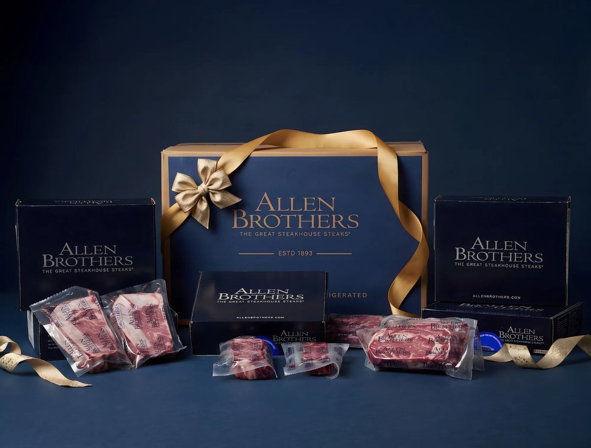 Allen Brothers gift set featuring a large navy blue and gold gift box with a ribbon, surrounded by vacuum-sealed raw steaks and smaller branded boxes on a dark blue background.