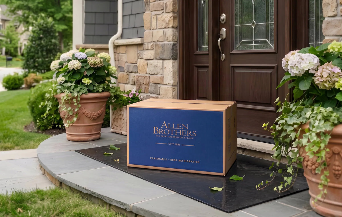 A wooden cooler storage box with a navy blue front panel and gold lettering reading 'Allen Brothers' sits on a black doormat at a front porch. The porch has stone walls, a dark wood door, and potted flowers on both sides.